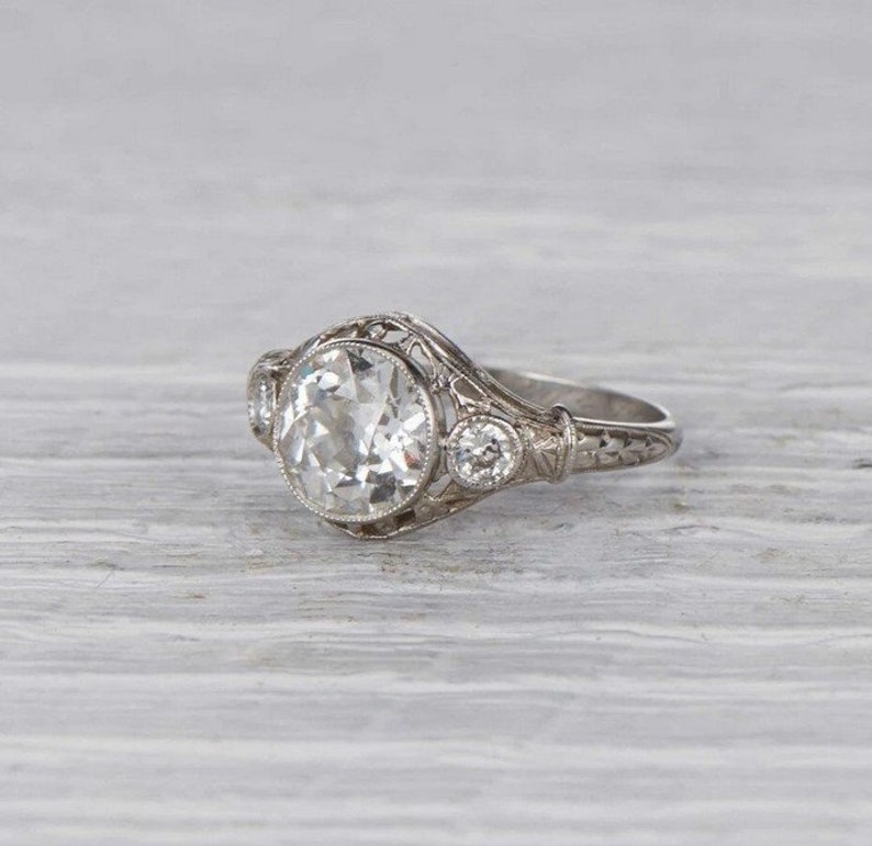 May include: A vintage engagement ring with a large, round, clear center stone and two smaller, round, clear accent stones. The ring is set in a delicate filigree design.