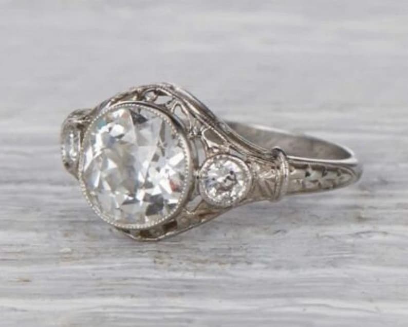 May include: A vintage-style engagement ring with a large, round, clear center stone surrounded by smaller, clear stones. The ring is made of white metal and has a delicate, filigree design.