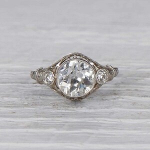 May include: A vintage engagement ring with a large, round, clear center stone and two smaller, round, clear accent stones. The ring is set in a delicate, filigree design.
