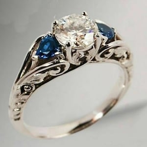 May include: A silver ring with a large, round, clear center stone and two smaller, teardrop-shaped, blue stones on either side.