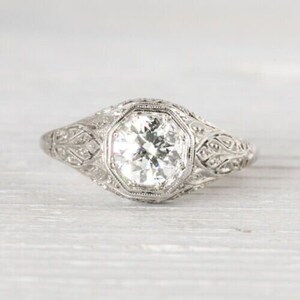 May include: A vintage-style engagement ring with a round, clear diamond set in a platinum band. The band is decorated with intricate filigree details.