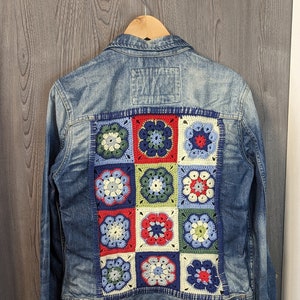 Könnte beinhalten: Eine blaue Jeansjacke mit einem gehäkelten Patch auf der Rückseite. Der Patch besteht aus neun Quadraten, jedes mit einem anderen floralen Design in Blautönen, Rot, Grün und Weiß.