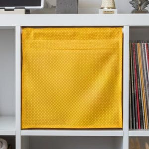 Puede incluir: Estantería cúbica blanca con varios objetos. Incluye un contenedor de almacenamiento de tela amarilla, una cesta de mimbre, libros, arte enmarcado, una tableta, auriculares y objetos decorativos. La estantería está contra una pared gris claro.