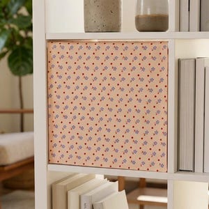 May include: A square fabric storage bin with a light peach background featuring a repeating pattern of small blue flowers and tiny red dots, positioned on a white shelving unit alongside books, a small ceramic vase, and a potted snake plant.