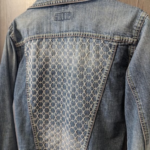 Può includere: Giacca di jeans blu chiaro con un motivo geometrico bianco ricamato sul retro. La giacca ha un colletto classico, chiusure con bottoni e una piccola toppa con logo sulla parte superiore della schiena.