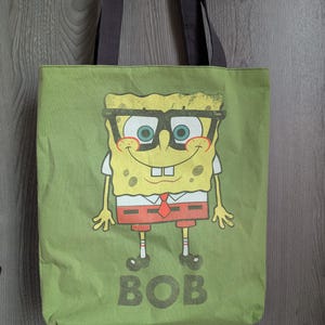 May include: Green canvas tote bag featuring a cartoon character with glasses, a white shirt, red tie, and orange shorts. The word "BOB" is printed in large, dark letters below the character. The bag has brown handles.