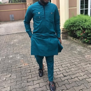 May include: Teal two-piece suit, comprising a long-sleeved tunic and trousers. The tunic has a small pocket and decorative button. The suit is worn with black loafers with gold detailing. A smart outfit suitable for formal events.