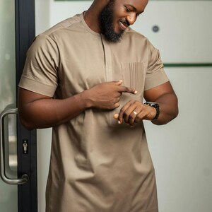 May include: A tan short-sleeved shirt with a pocket on the chest. The shirt has a textured pattern and a mandarin collar. The person is wearing a black watch and a ring. The outfit is part of a formal wear collection.