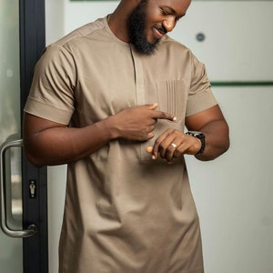 May include: A tan short-sleeved shirt with a pocket on the chest. The shirt has a textured pattern and a mandarin collar. The person is wearing a black watch and a ring. The outfit is part of a formal wear collection.