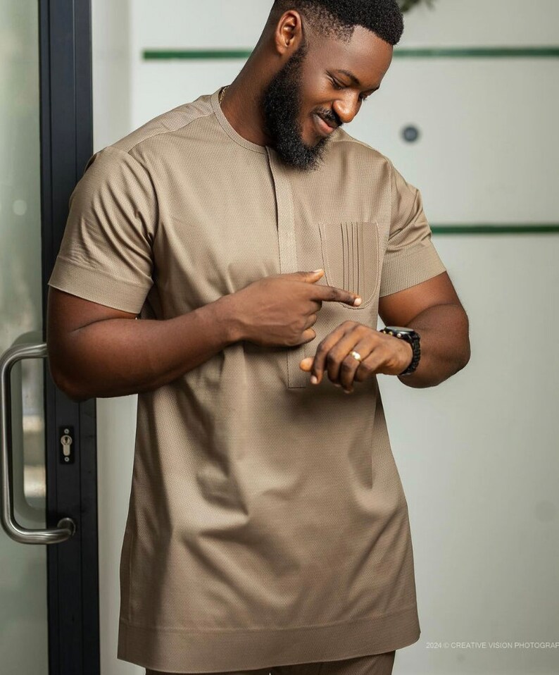 May include: A tan short-sleeved shirt with a pocket on the chest. The shirt has a textured pattern and a mandarin collar. The person is wearing a black watch and a ring. The outfit is part of a formal wear collection.