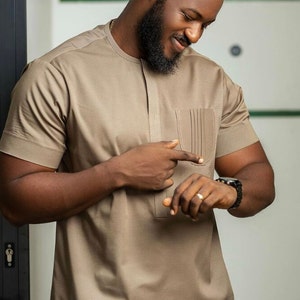 May include: A tan short-sleeved shirt with a textured pattern and a pocket on the chest. The shirt has a mandarin collar and a buttoned placket. The person is wearing a black watch with a black band.