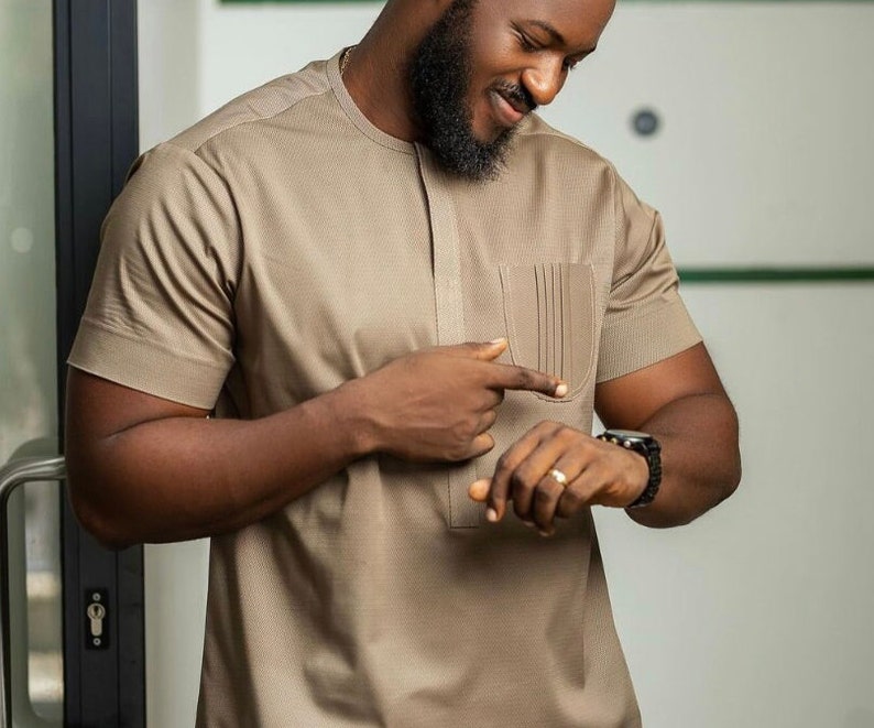 May include: A tan short-sleeved shirt with a textured pattern and a pocket on the chest. The shirt has a mandarin collar and a buttoned placket. The person is wearing a black watch with a black band.