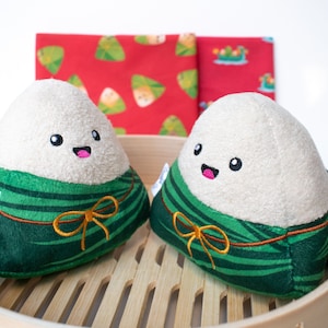 May include: Two green and white plush rice dumplings with happy faces and gold bows. The dumplings are sitting in a wooden steamer basket. The text "Cookies & Co." is on the left dumpling.