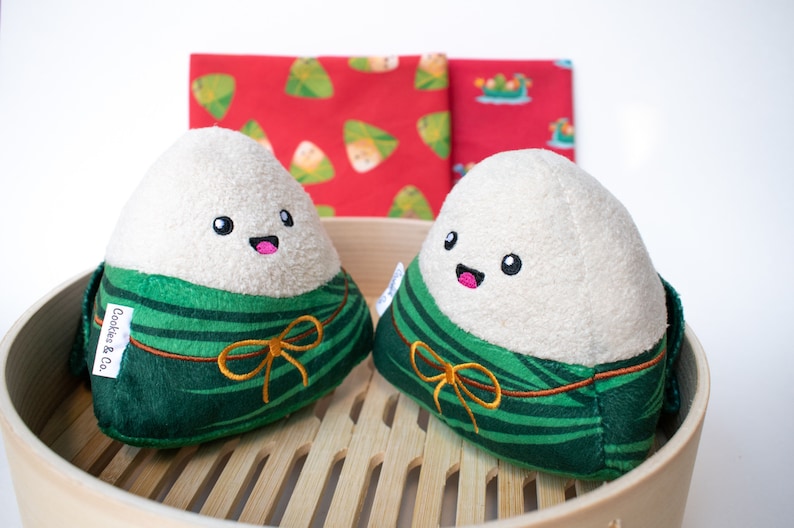 May include: Two green and white plush rice dumplings with happy faces and gold bows. The dumplings are sitting in a wooden steamer basket. The text "Cookies & Co." is on the left dumpling.