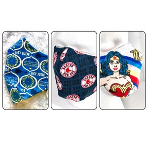 May include: Three fabric face masks. The first mask is blue with a white and yellow pattern and the text "Hey Jude" repeated. The second mask is dark blue with a white and red pattern of the Boston Red Sox logo. The third mask is white with a red, yellow, and blue Wonder Woman graphic.