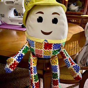 May include: A plush doll with a white head, black eyes, and a smiling mouth. It wears a yellow hat with "BoBo Jr" printed on it. The doll's body and limbs are made of colorful patchwork fabric.