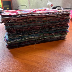 May include: A tall, thick stack of square fabric pieces layered neatly on top of each other. The top piece features a vibrant red background with a repeating pattern of colorful, stylized floral and geometric motifs in shades of blue, yellow, and white.