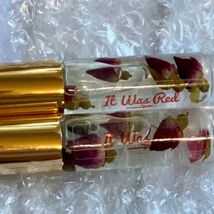 May include: Two clear glass roller bottles with gold lids. Each bottle contains dried pink rose petals and clear liquid. The bottles are labeled "It Was Red" with a red arrow.