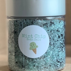 May include: A clear jar filled with green and black bath salts. The jar has a silver lid and a white label that says "Mint Chip Bath Salt" with a drawing of a mint chip ice cream cone.