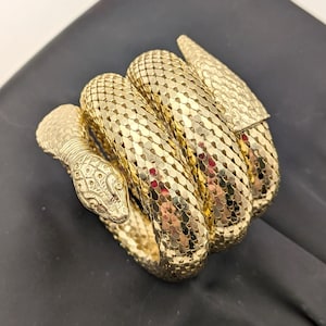 May include: A gold-toned metal bracelet shaped like a coiled snake. The surface features a textured, scale-like pattern throughout the entire body, with detailed engravings on the head and a smooth, tapered tail end that wraps around the wrist.