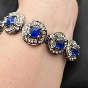 May include: A silver-toned bracelet featuring square, deep blue gemstones at the center of each circular link. The links are surrounded by smaller, clear crystals. The bracelet is shown on a wrist.