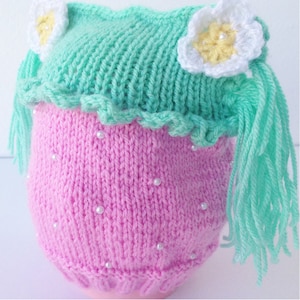 Può includere: Un completo per neonati lavorato a mano a maglia a forma di fragola. Il completo è principalmente rosa con una parte superiore verde e dettagli di fiori bianchi. Piccole perle bianche sono sparse sulla parte rosa. Il completo è progettato per assomigliare a una fragola.