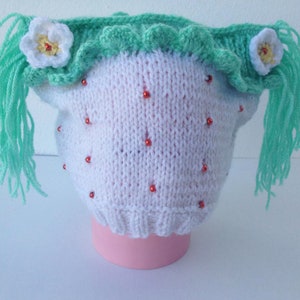 Può includere: Un cappello bianco lavorato a maglia a mano con dettagli di perline rosse, che ricorda una fragola. Il cappello presenta un bordo verde smerlato e due decorazioni floreali bianche con centri gialli. Lunghe ciocche di filato verde menta pendono dai lati.
