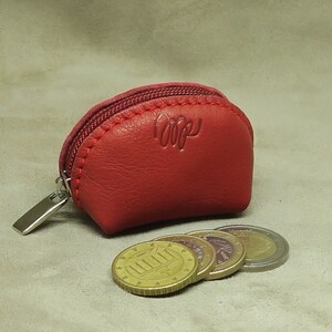 May include: A small, red leather coin purse with a zipper closure. The purse has the word "LOopy" embossed on the front. The purse is sitting on a beige surface with four euro coins in front of it.