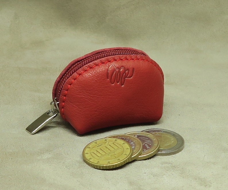 May include: A small, red leather coin purse with a zipper closure. The purse has the word "LOopy" embossed on the front. The purse is sitting on a beige surface with four euro coins in front of it.