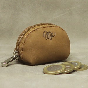 May include: A small, brown leather coin purse with a zipper closure. The purse has a stitched design and a small inscription on the front. The purse is sitting on a beige surface with several coins next to it.