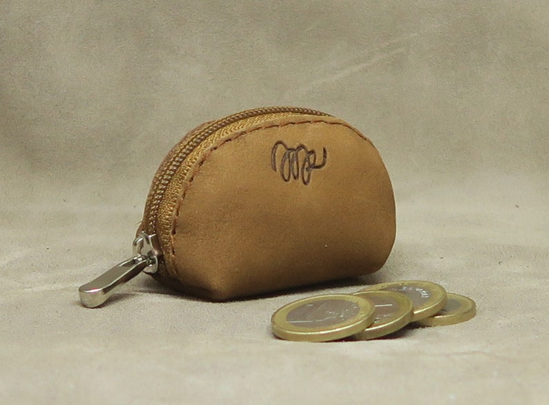 May include: A small, brown leather coin purse with a zipper closure. The purse has a stitched design and a small inscription on the front. The purse is sitting on a beige surface with several coins next to it.