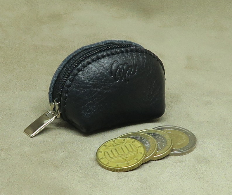 May include: A small black leather coin purse with a zipper closure and a silver zipper pull. The purse is sitting on a beige surface with four gold coins next to it.