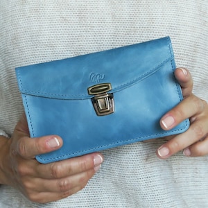 May include: A light blue leather wallet with a flap closure and a brass-colored clasp. The wallet has a stitched border and a small logo on the top flap. The wallet is held in two hands.
