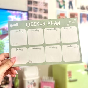 May include: A light green weekly planner with the words "WEEKLY PLAN" in white. The planner has boxes for each day of the week, with cute paw prints and bone illustrations. The planner is held by a person.