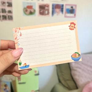 May include: A lined, rectangular notepad with a peach border, featuring small illustrations of a boat, a bowl of ramen, and a character. The notepad is held in a hand, with other images in the background.
