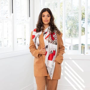 以下が含まれることがあります： 茶色のブレザーとそれに合わせたパンツを着用した女性が、白と赤の模様の入ったスカーフをしています。スカーフは、黒い線と赤いアクセントがついた幾何学模様です。