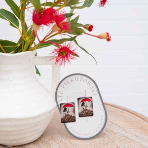 May include: A pair of square wooden earrings with a colorful illustration of a woman walking under a red umbrella. The earrings are on a white card with the text "ATE PIEKUTOWSKI CONTEMPORARY PRINTMAKER".