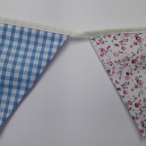 May include: A blue and white gingham fabric bunting flag with a white floral fabric bunting flag. Both flags are sewn with a white trim.