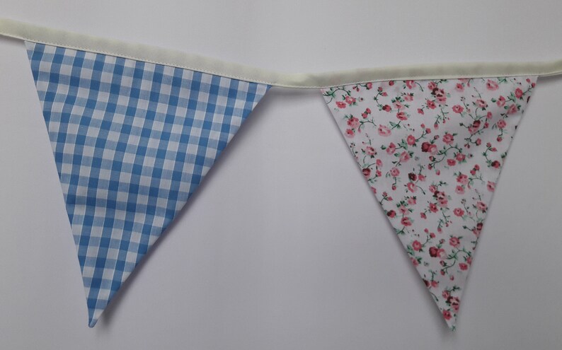 May include: A blue and white gingham fabric bunting flag with a white floral fabric bunting flag. Both flags are sewn with a white trim.
