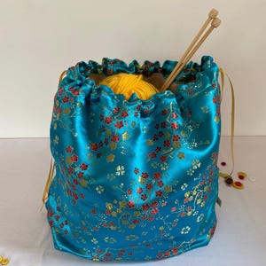 Puede incluir: Una bolsa de satén turquesa con cordón y un estampado floral en rojo y dorado. La bolsa contiene un ovillo de lana amarilla y dos agujas de tejer de madera. La bolsa está atada con una cinta dorada.