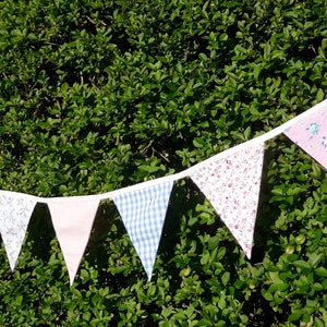May include: A bunting with a white background and colorful fabric triangles. The triangles feature floral patterns in pink, blue, and white. The bunting is strung on a white cord and is hanging against a green hedge.