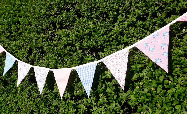 May include: A bunting with a white background and colorful fabric triangles. The triangles feature floral patterns in pink, blue, and white. The bunting is strung on a white cord and is hanging against a green hedge.