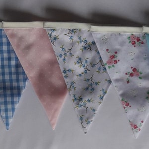 May include: A bunting banner with 10 triangular flags in a variety of pastel colors and patterns, including pink, blue, and white floral prints, gingham, and polka dots.