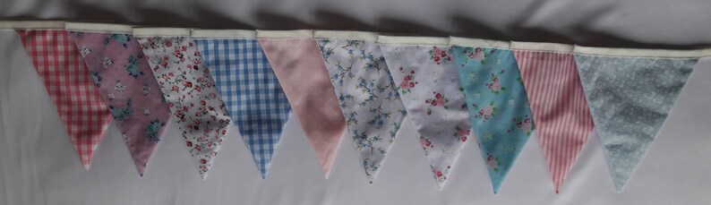 May include: A bunting banner with 10 triangular flags in a variety of pastel colors and patterns, including pink, blue, and white floral prints, gingham, and polka dots.