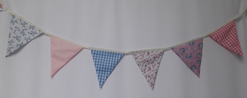 May include: A bunting banner with seven triangular flags. The flags are made of fabric and feature different patterns, including floral, gingham, and polka dots. The flags are connected by a white string.