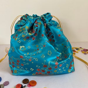 May include: A turquoise blue drawstring bag with a floral pattern in gold and red. The bag has a gold drawstring and is made of a silky fabric.