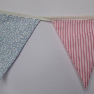 May include: A bunting with alternating blue floral and pink striped fabric triangles. The bunting is strung together with white ribbon.