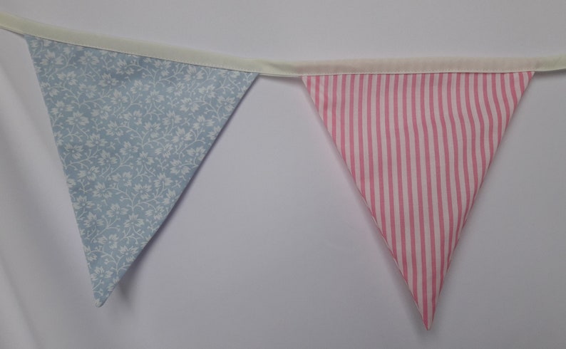 May include: A bunting with alternating blue floral and pink striped fabric triangles. The bunting is strung together with white ribbon.