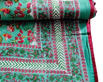 May include: A rolled-up, turquoise-colored quilt with a floral pattern in red and green. The quilt has a border with a chevron design in white and green, and a red border. The floral pattern includes flowers and leaves.