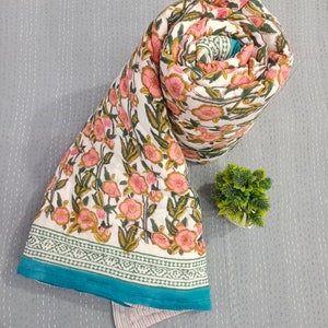 May include: A white cotton quilt with a floral print in shades of pink and green. The quilt is rolled up and has a teal border with a green and white patterned trim.